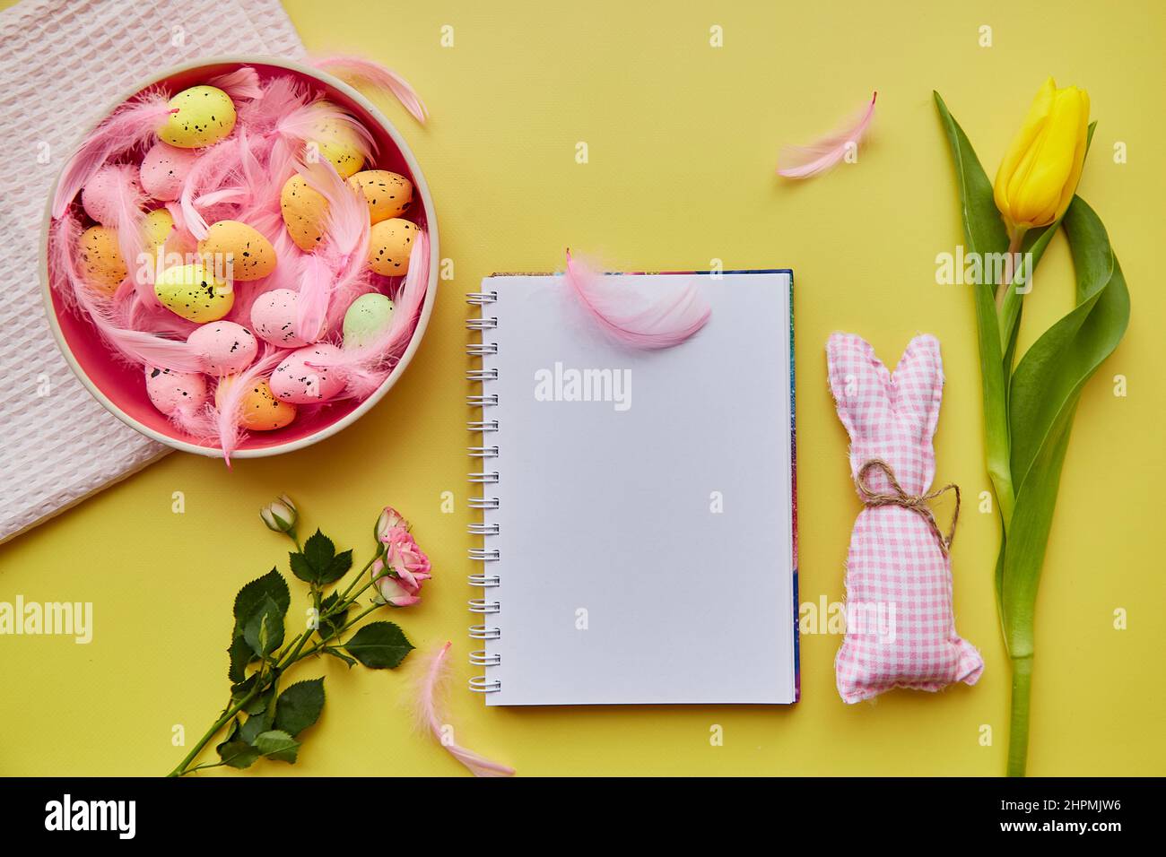 Easter-Notizblock im Nachbau. Rosa Schale mit bunten Eiern, rosa Federn, basteln niedlichen Hasen und Blumen. Happy Easter Konzept. Draufsicht. Postkartenmock auf gelbem Hintergrund Stockfoto