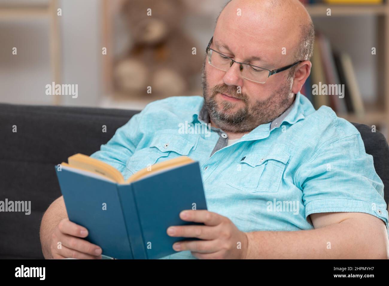 Ein kahler Mann, der auf einem Sofa sitzt, liest zu Hause ein Buch. Stockfoto