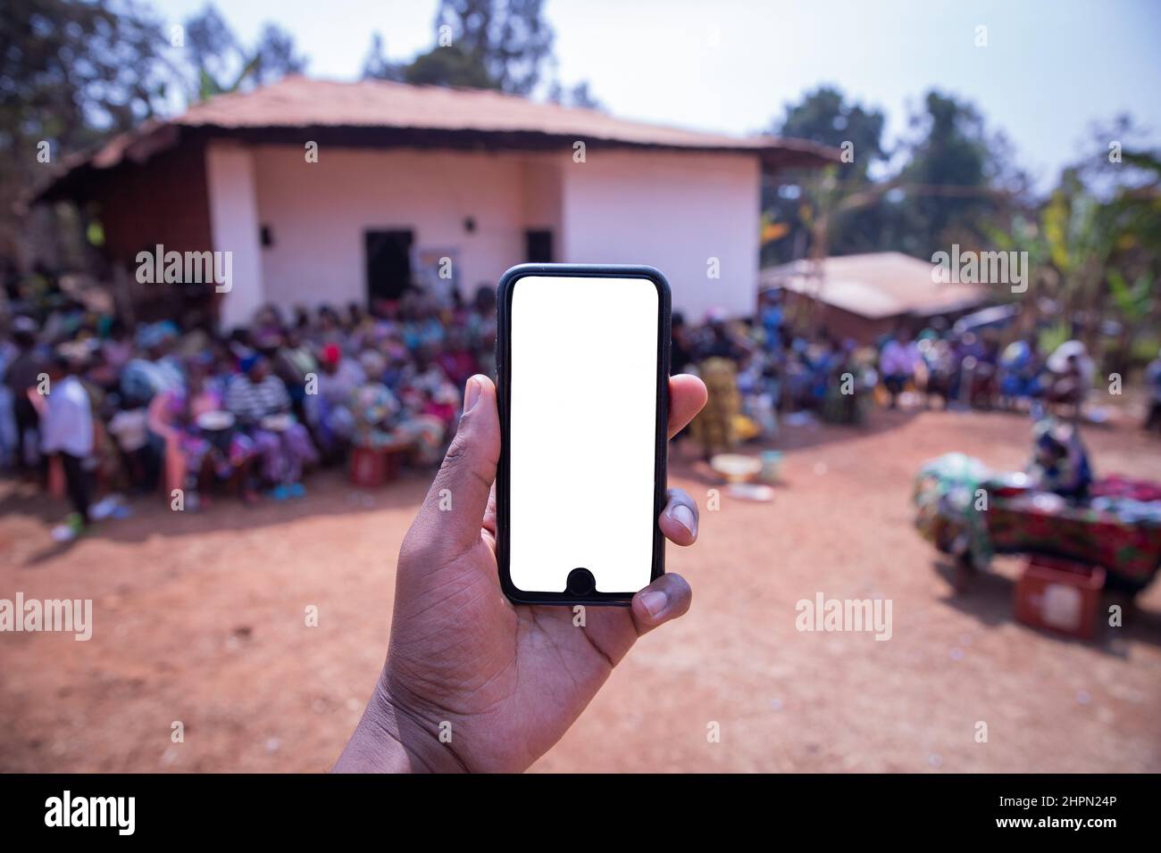 Modell des Bildschirms eines Smartphones, das von einer Hand gehalten wird, mit einem afrikanischen Dorf im Hintergrund, in dem Menschen saßen Stockfoto