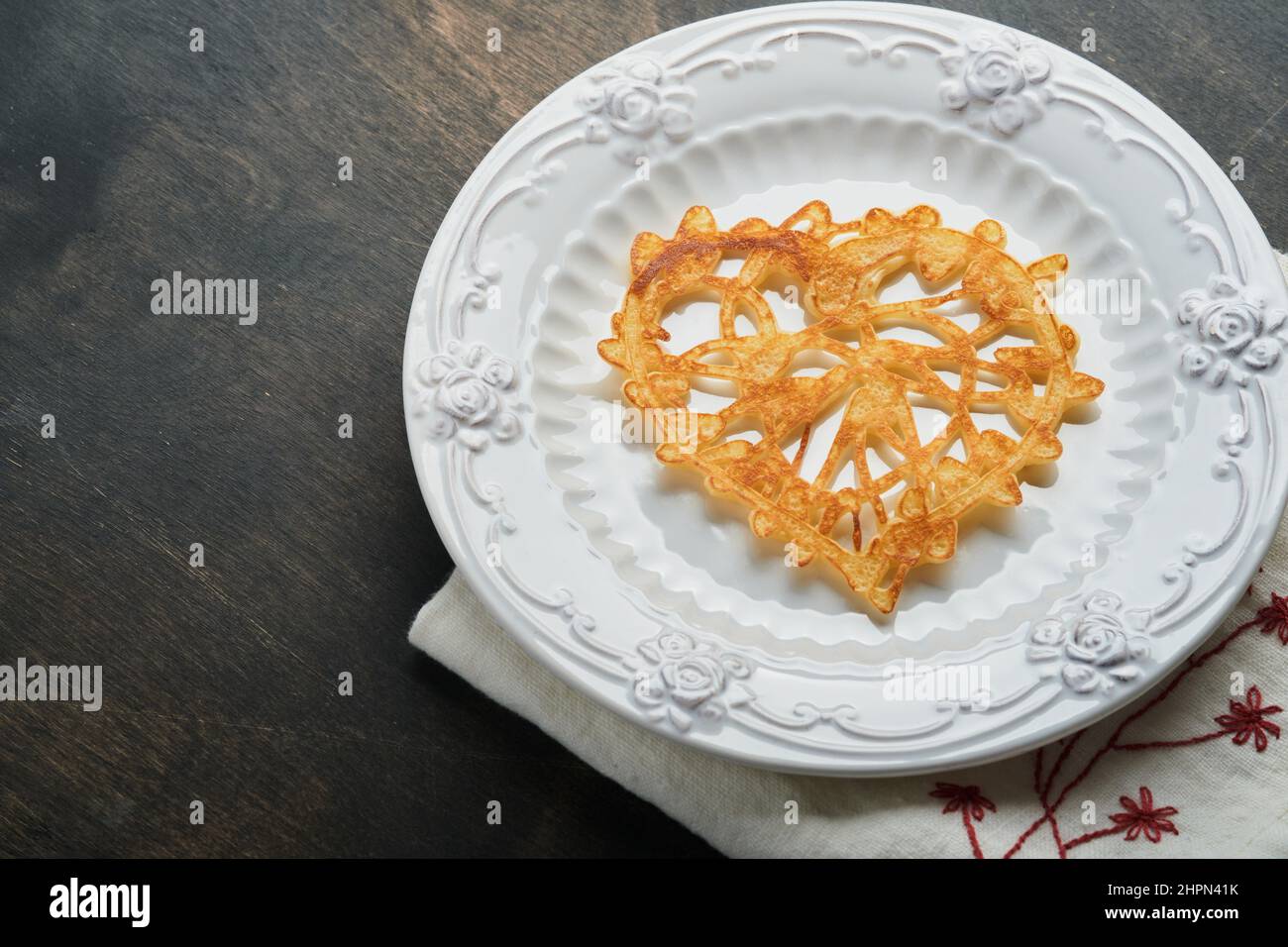 Herzförmige Spitzenpfannkuchen oder Crepes auf weißem Teller auf altem rustikalem Hintergrund. Tischeinstellung für Ihr Lieblingsfrühstück am Valentinstag. Kopie der Draufsicht Stockfoto