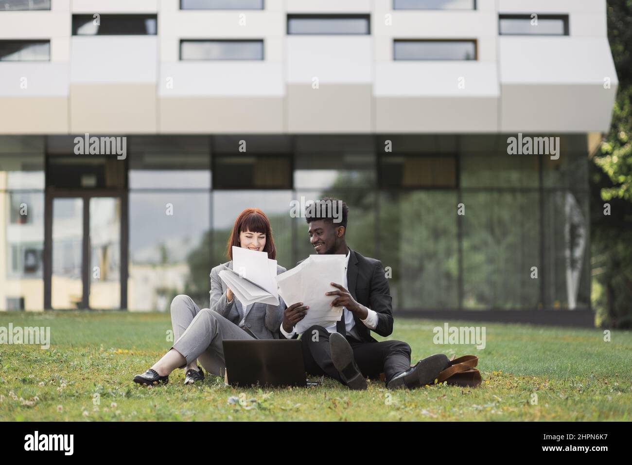 Zwei freudig aufgeregt selbstbewusste multiethnische Geschäftsleute, sitzen vor dem Büro auf dem Rasen und lesen Papiere. Glückliche Kollegen sitzen mit Papieren auf dem Rasen und sprechen über das Projekt Stockfoto