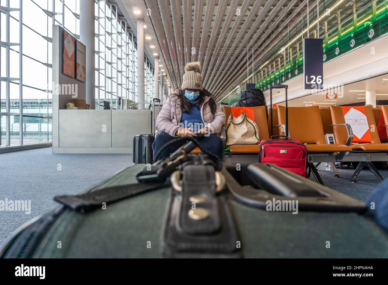 Flughafen Charles de Gaulle, Paris - eine weibliche ethnische Passagierin mit Gesichtsmaske und Checks auf ihrem Telefon, mit einem Koffer im Vordergrund Stockfoto