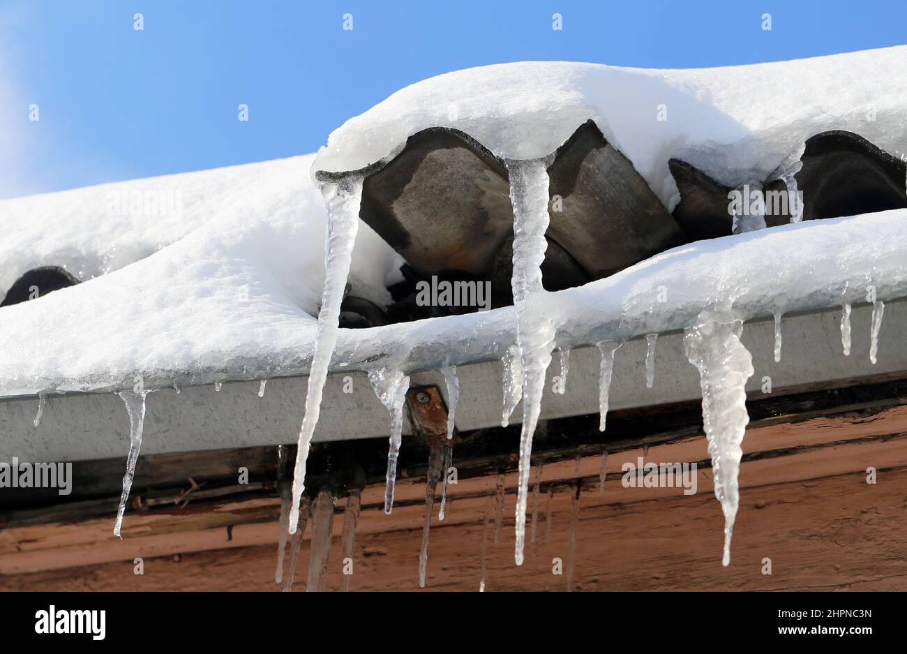 Eiszapfen hängen auf dem schneebedeckten Dach des Hauses gegen den blauen Himmel Stockfoto
