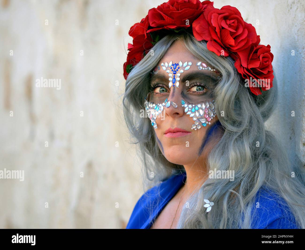 Junge, schöne kaukasische Frau mit roten Blumen in ihren grauen Haaren und vielen Gesichtssteinen am mexikanischen Tag der Toten (Día de los Muertos) sieht den Betrachter an. Stockfoto