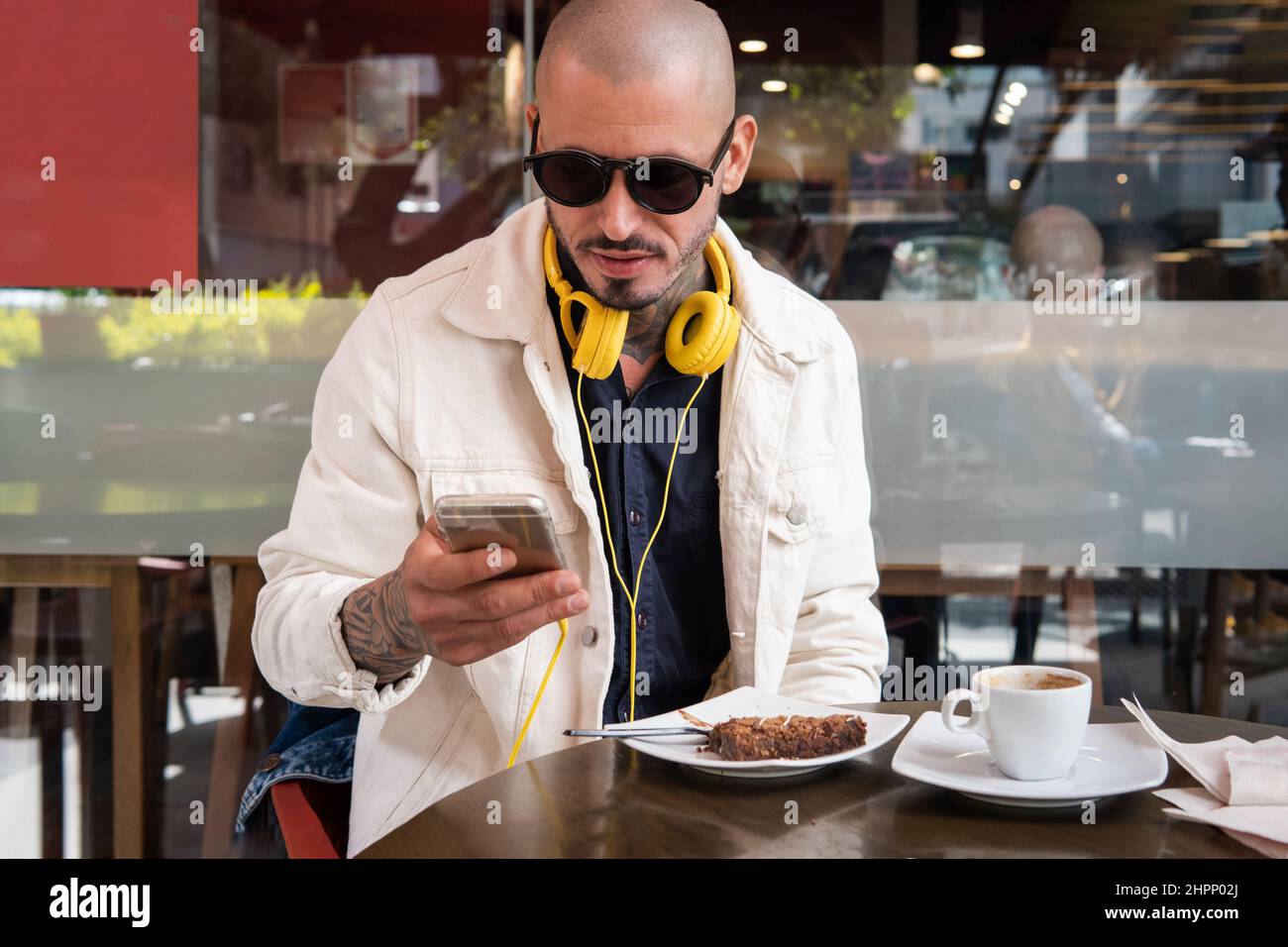 Ein junger Mann mit gelben Kopfhörern plaudert in einem Café mit seinem Handy, trinkt Kaffee und isst einen Kuchen Stockfoto
