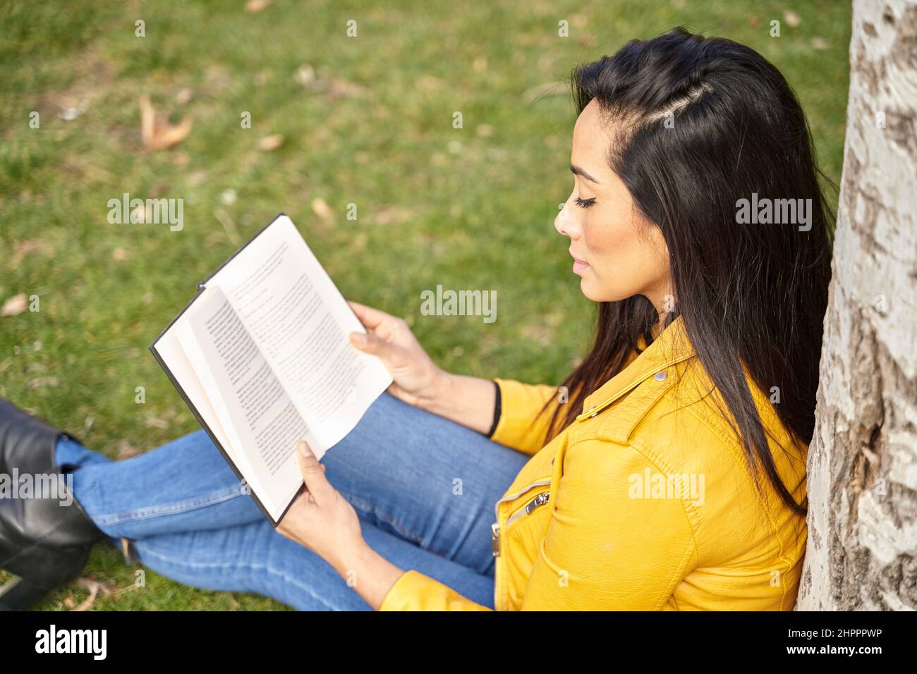 Seitenansicht einer nachdenklichen hispanischen Frau beim Lesen eines Romans, während sie mit einem Buch unter einem Baum im Sommergarten saß Stockfoto