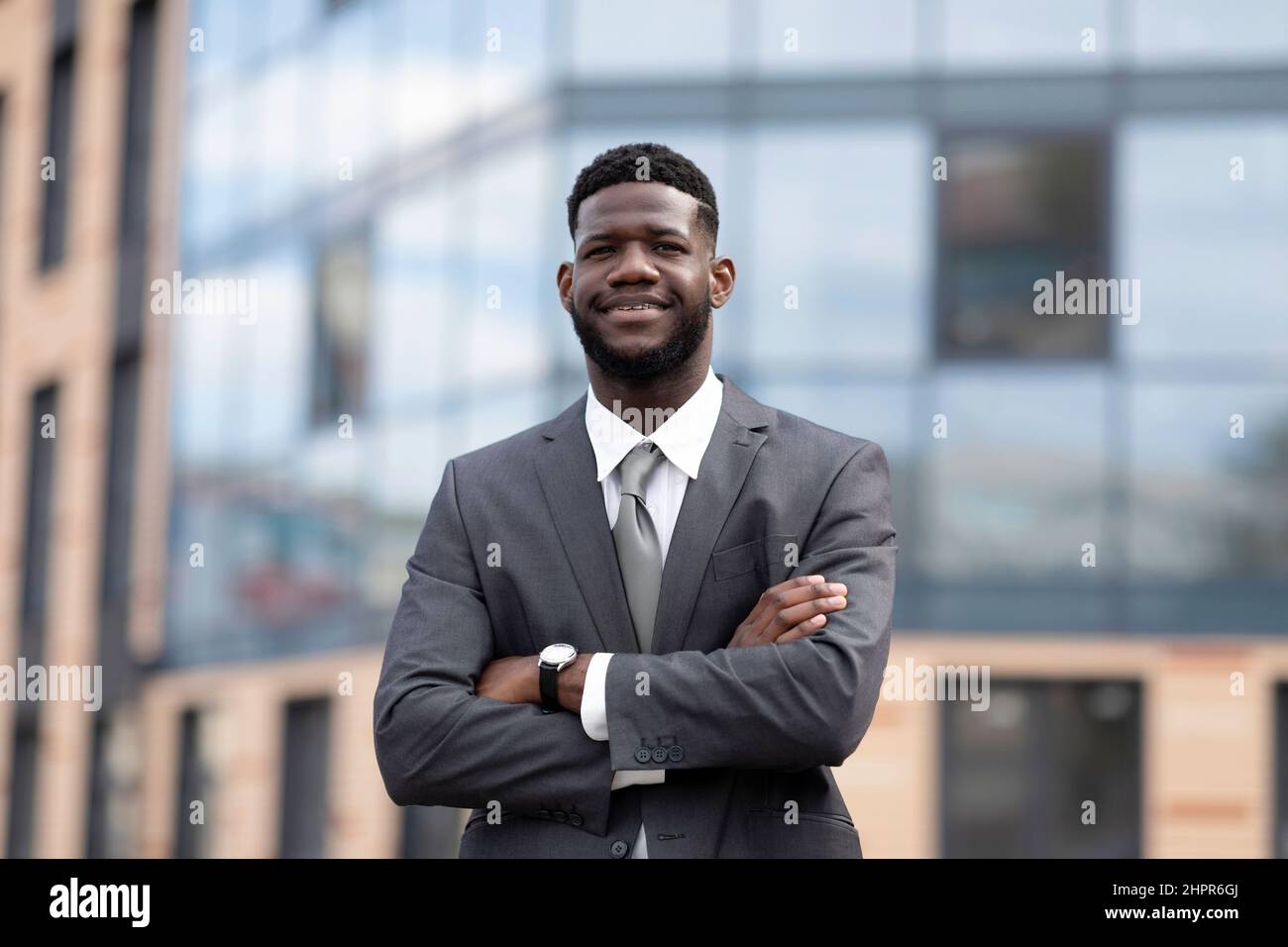 Wohlhabender afroamerikanischer Unternehmer, junger Mann, der vor dem modernen Bürogebäude steht und mit gefalteten Armen posiert Stockfoto