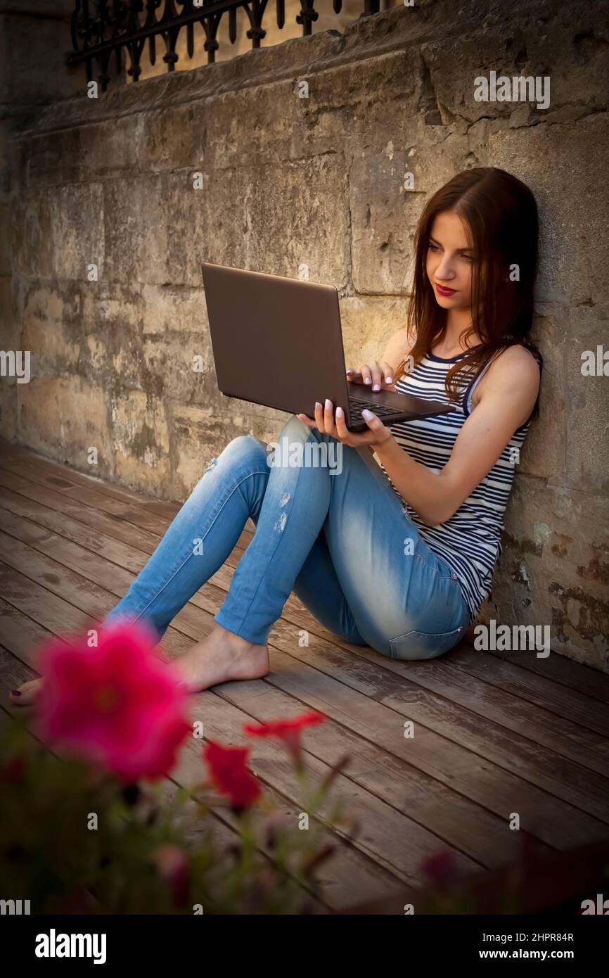 Schöne junge Geschäftsfrau freiberufliche Eingabe auf Laptop im Freien auf einem Holzboden. Die besten Orte, an denen man in einer Stadt Remote arbeiten kann. Vertikales Bild. Stockfoto