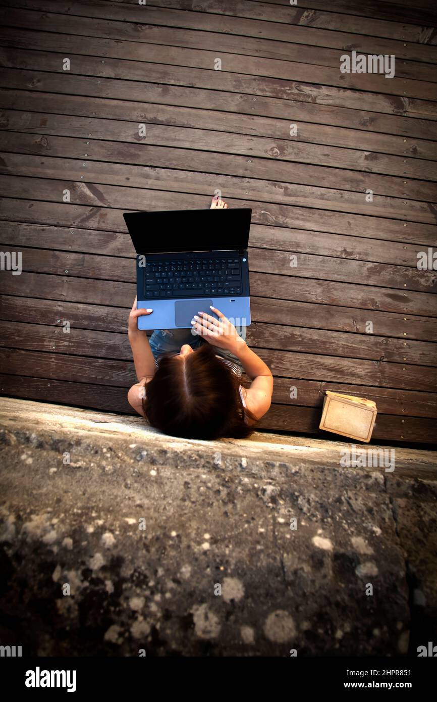 Draufsicht auf die schöne junge Geschäftsfrau, die freiberuflich auf einem Laptop im Freien auf einem Holzboden tippt. Die besten Orte, an denen man in einer Stadt Remote arbeiten kann. Vertikale ima Stockfoto