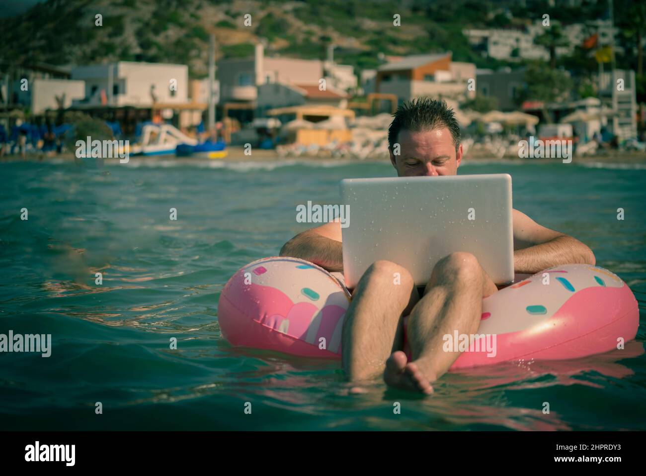 Freiberufler mit einem aufblasbaren Ring im Wasser des Meeres arbeitet am Computer. Glücklicher Mann im Schwimmkreis mit Beinen im Wasser und mit silbernem Laptop. Stockfoto