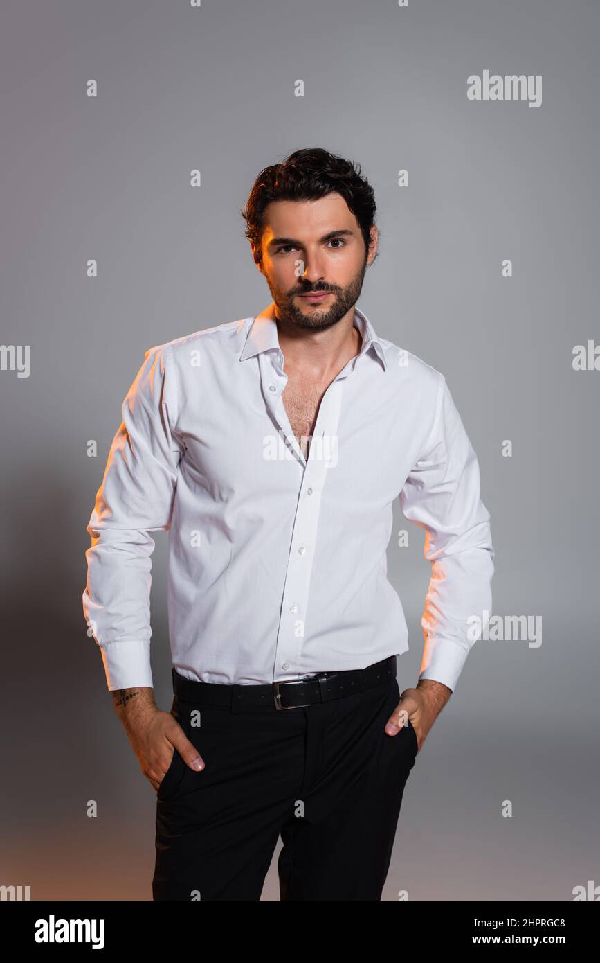 Stilvoller Mann im weißen Hemd, der mit Händen in schwarzen Hosentaschen auf grau steht Stockfoto