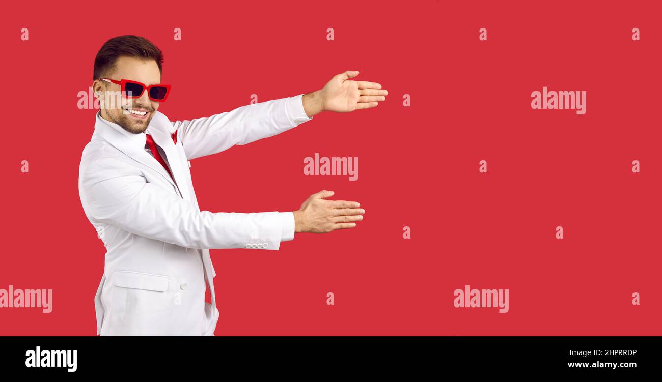 Hübscher Mann in weißem Anzug und Sonnenbrille, der etwas auf rotem Bannerhintergrund zeigt Stockfoto