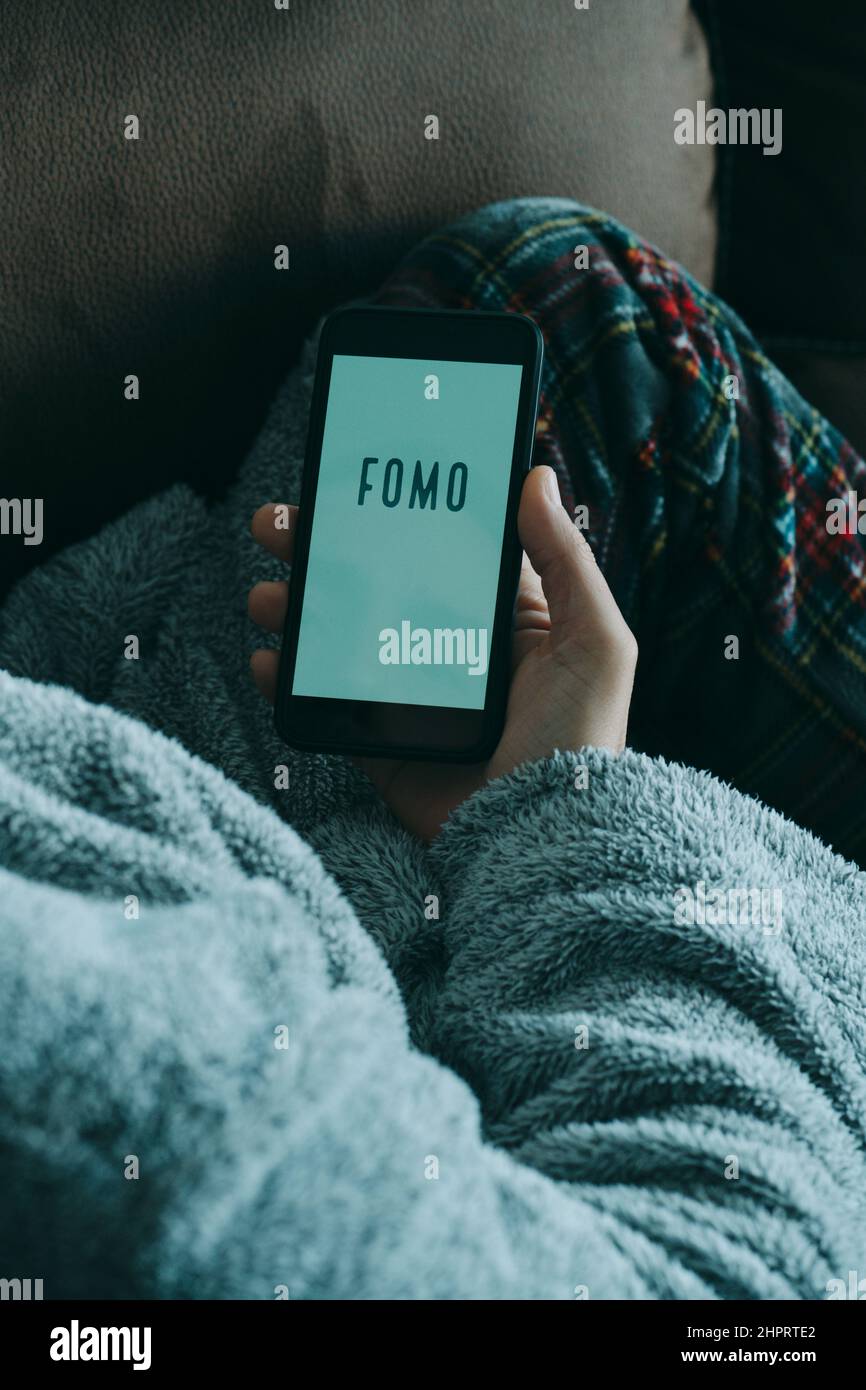 Nahaufnahme eines jungen kaukasischen Mannes, der Schlafanzug und Hausmantel trägt, auf dem Sofa liegt und sein Smartphone beobachtet, das den Text fomo liest, aus Angst vor Missi Stockfoto