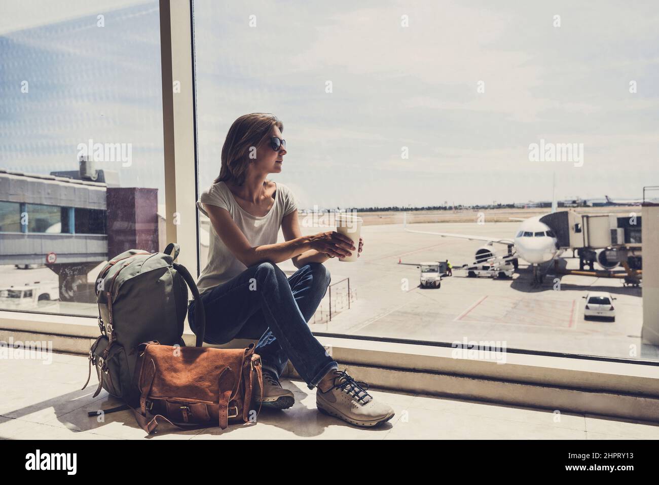 Junge Frau am Flughafen, die durch das Fenster auf Flugzeuge schaut und Kaffee trinkt, Reisen, Urlaub und aktives Lifestyle-Konzept Stockfoto