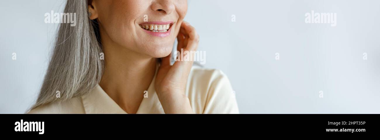 Attraktive silberhaarige Frau in beigem Hemd berührt Wange auf hellgrauem Hintergrund Stockfoto