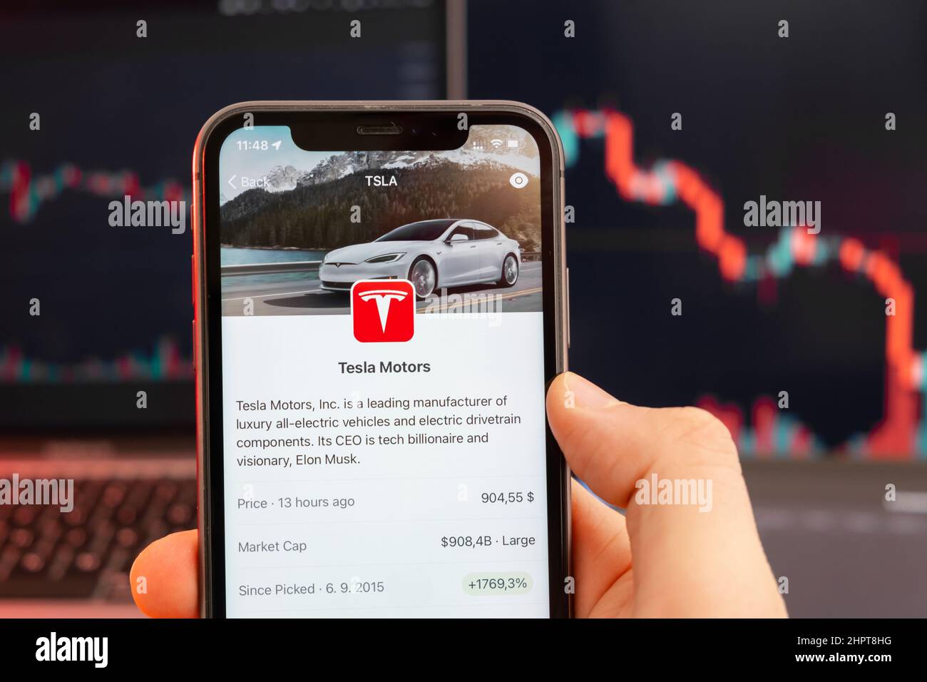 Abwärtstrend der Aktienkursen von Tesla Motors mit roten Grafiken im Hintergrund. Man hands Holding a Smartphone with Logo on the screen, February 2022, San Francisco, USA. Stockfoto