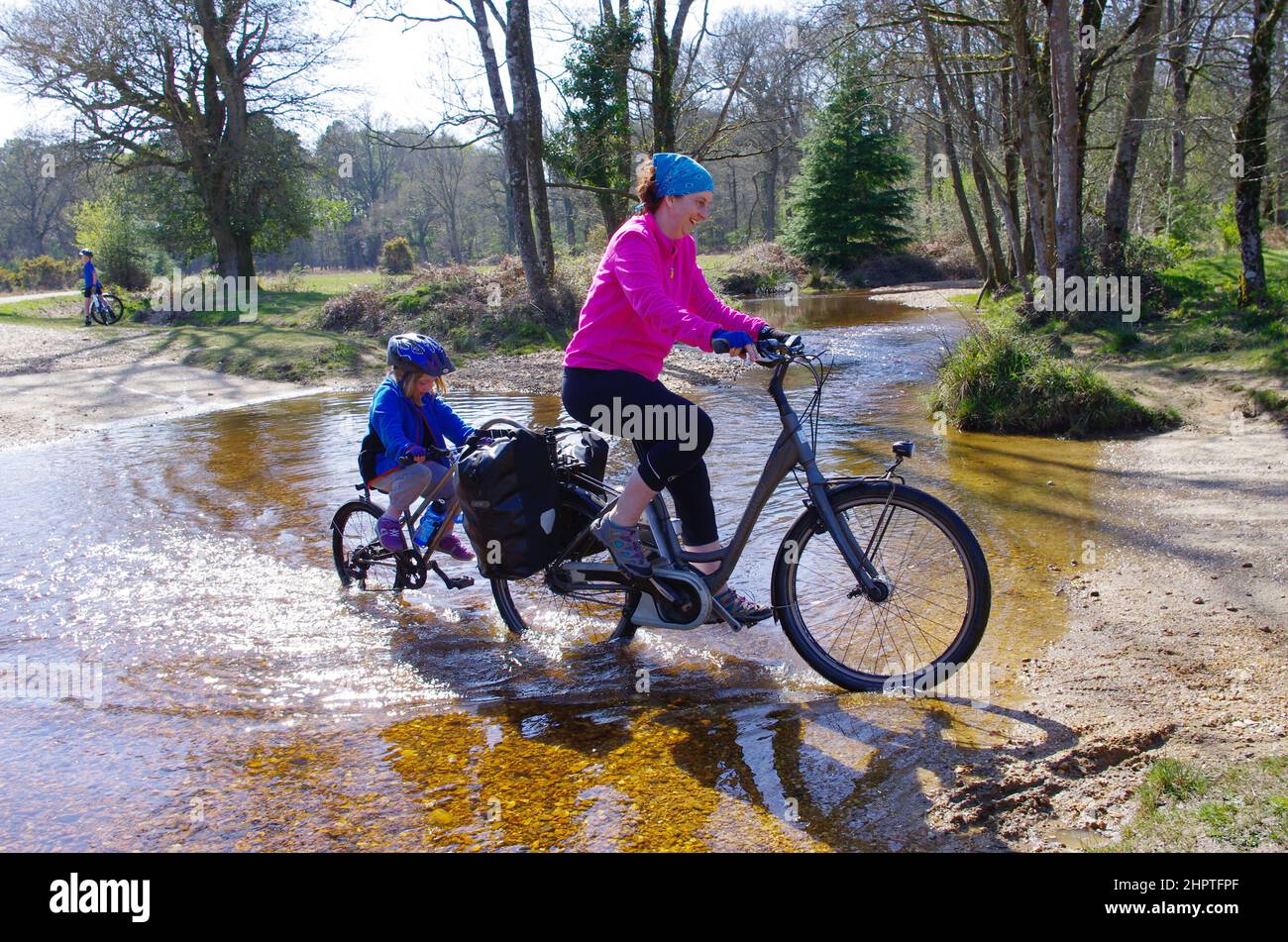 Frau auf einem Elektrofahrrad und Kind auf einem Tagalong (Anhängerfahrrad), das durch Fletchers Water ford im New Forest National Park fährt Stockfoto