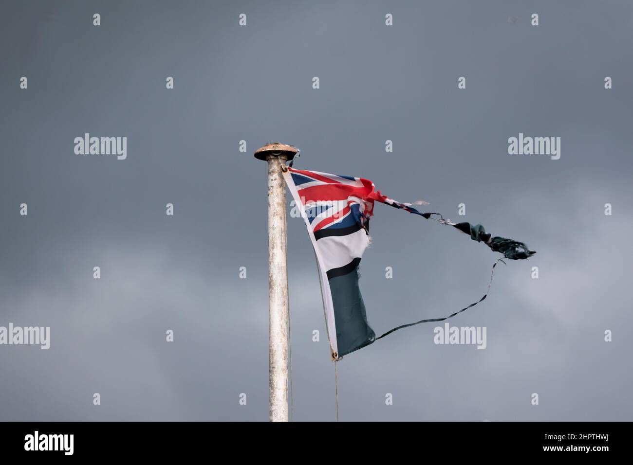 Eine zerrissene Union Jack-Flagge fliegt an einem launischen Tag von einem weißen Fahnenmast. Dunkle Wolken tragen zum düsteren Bild bei. Stockfoto