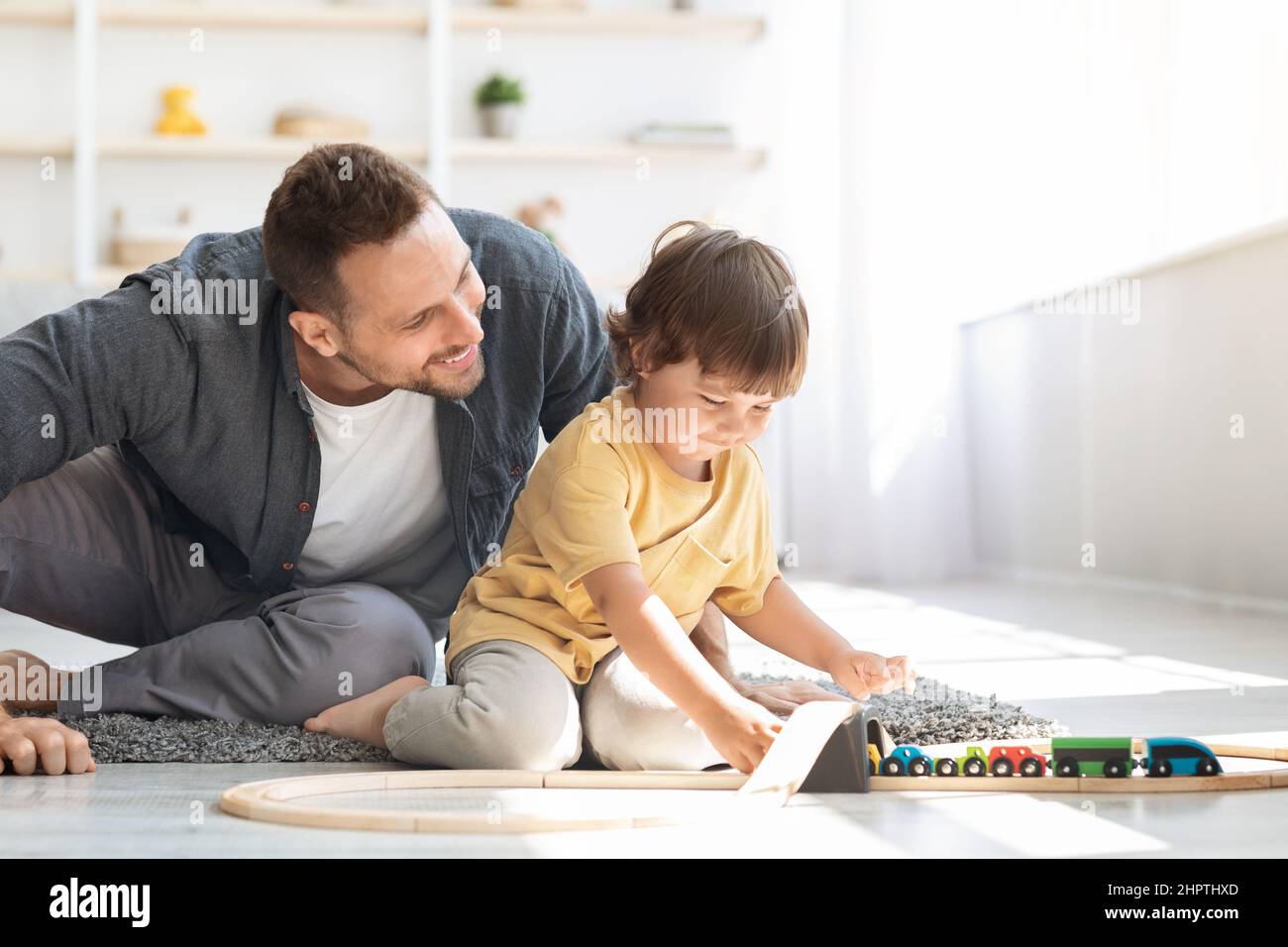 Niedlicher kleiner Junge, der mit einem liebevollen Vater einen Holzzug und eine Eisenbahn spielt und zu Hause auf dem Boden sitzt, leerer Platz Stockfoto