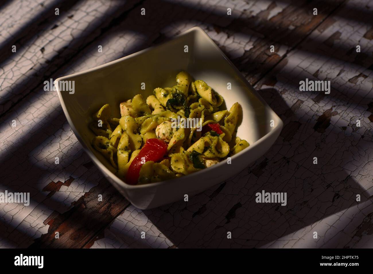 Mozzarella, Tomaten und Pesto Nudelsalat in einer weißen quadratischen Schale auf einem verzweifelten Tisch mit Sonnenfensterschatten, die über den Tisch und das Gericht fallen. Stockfoto