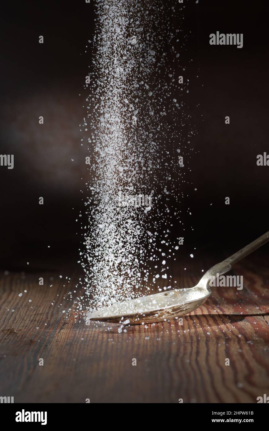 Ein Salzfall auf dunklem Grund, ein Löffel Salz liegt darauf. Grobes Salz Stockfoto