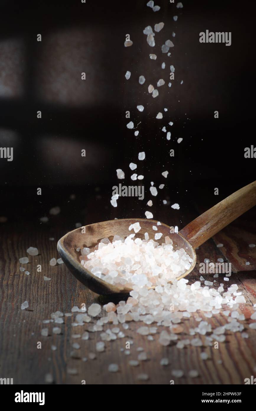 Ein Salzfall auf dunklem Grund, ein Löffel Salz liegt darauf. Grobes Salz Stockfoto
