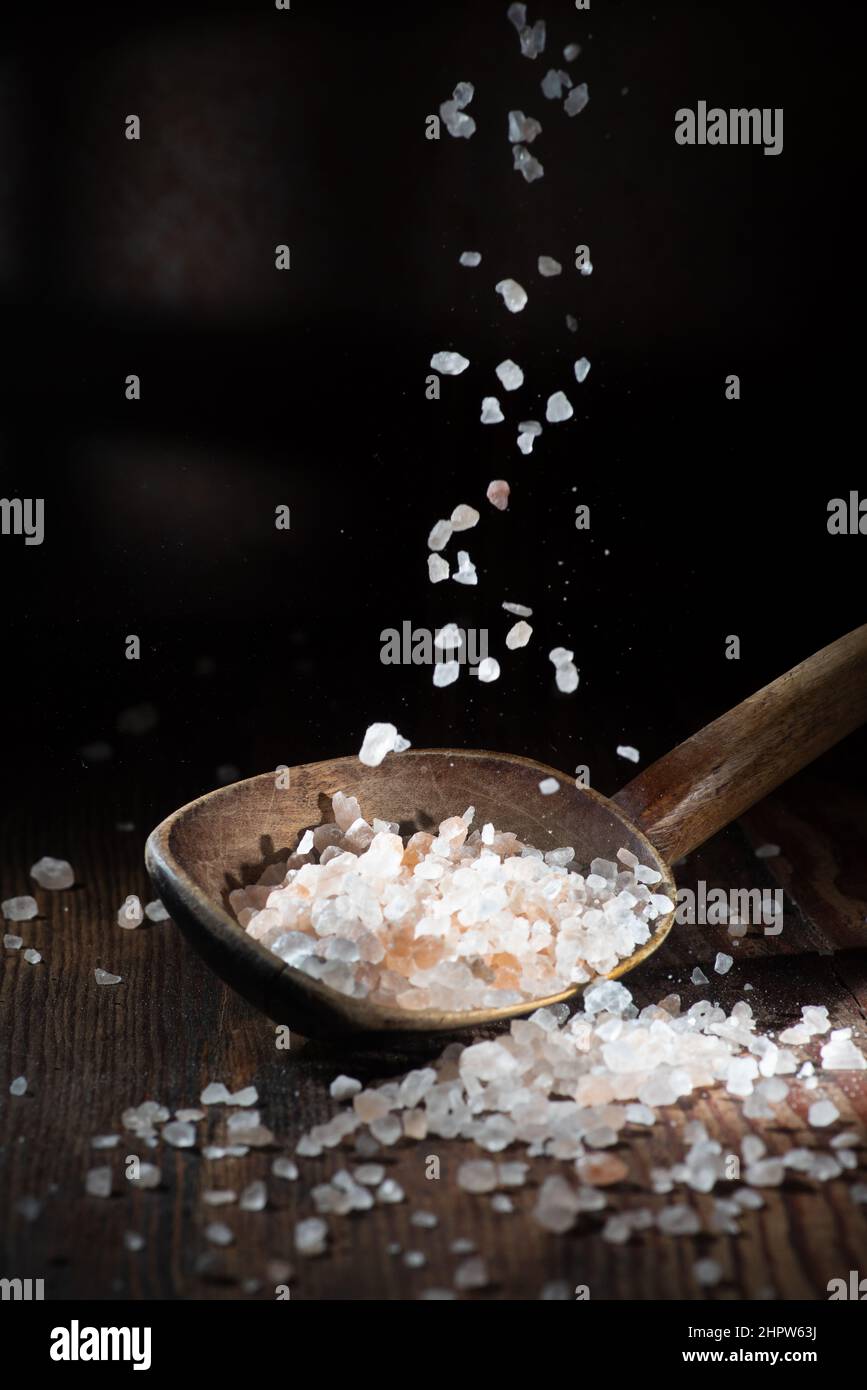 Ein Salzfall auf dunklem Grund, ein Löffel Salz liegt darauf. Grobes Salz Stockfoto