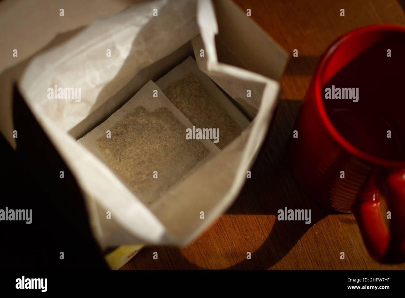 Eine offene Kiste mit Teebeuteln und einem roten Becher sitzt im goldenen Stundenlicht auf dem Holztisch. Stockfoto