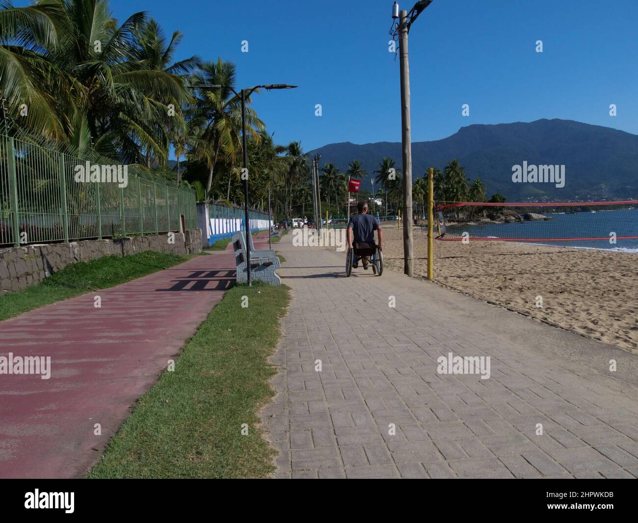 Ein behinderter Mann im Rollstuhl nähert sich dem Betrachter. Er nutzt den Fußgängerweg auf der Insel Ilhabela, Brasilien, was sehr praktisch ist Stockfoto