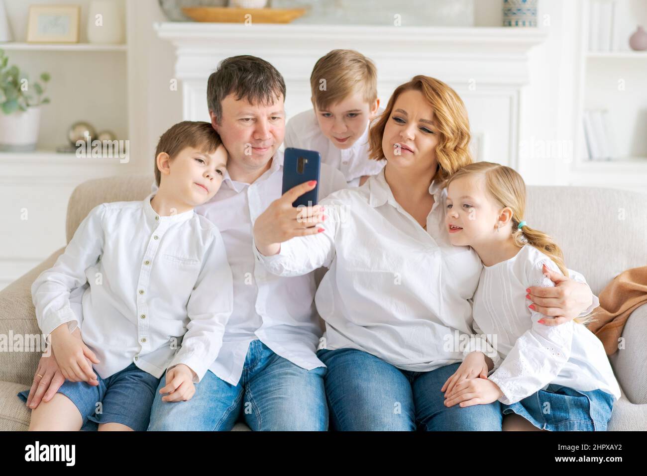 Kaukasische Familie sitzt auf dem Sofa im Wohnzimmer zu Hause. Mutter Vater kleine Tochter und zwei Söhne, die ein Smartphone benutzen, schauen sich ein Unterhaltungsprogramm an, Videos online und sprechen bei einem Videoanruf Stockfoto