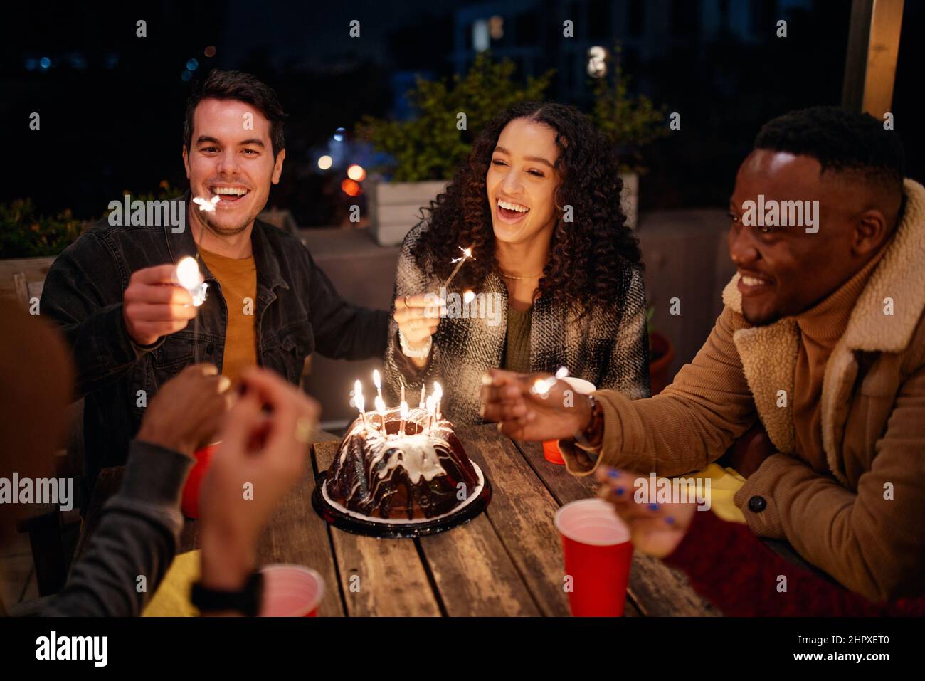 Eine Gruppe von jungen Erwachsenen, die auf der Geburtstagsfeier auf dem Dach mit Boho-Motto sitzen und Wunderkerzen aus Kuchen erleuchten. Stockfoto