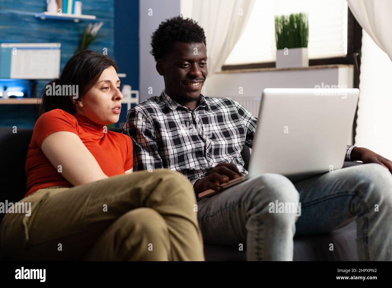 Gemischtrassig entspanntes Paar, das auf den Laptop-Bildschirm schaut, während es auf der Couch im Wohnzimmer sitzt. Junge Menschen genießen gemeinsam am Computer das Surfen im Internet und soziale Medien, während sie sich lustige Videos ansehen Stockfoto