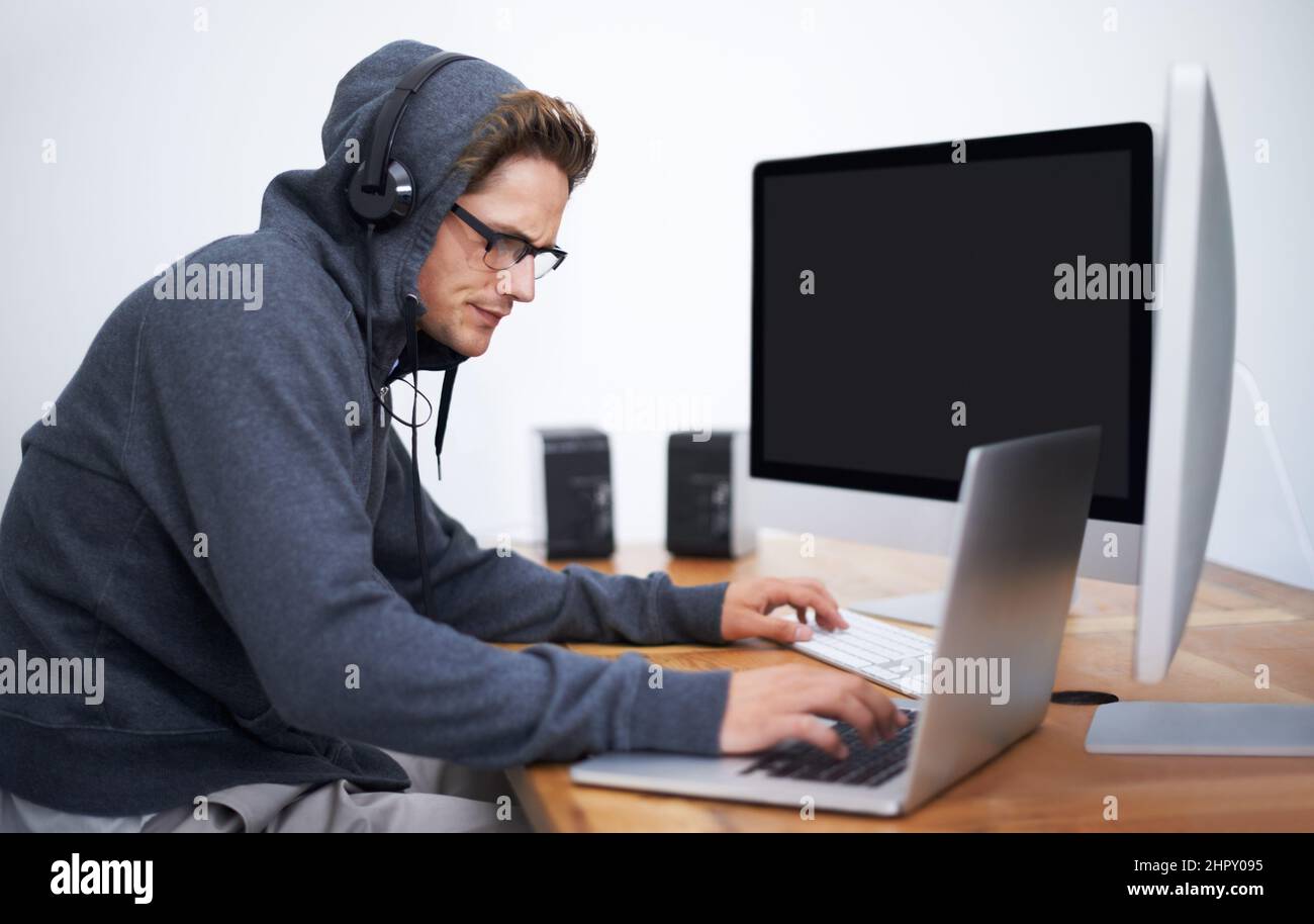 In seine Arbeit vertieft. Ein junger Hacker, der auf zwei Bildschirmen und einem Laptop mit Hoodie arbeitet. Stockfoto