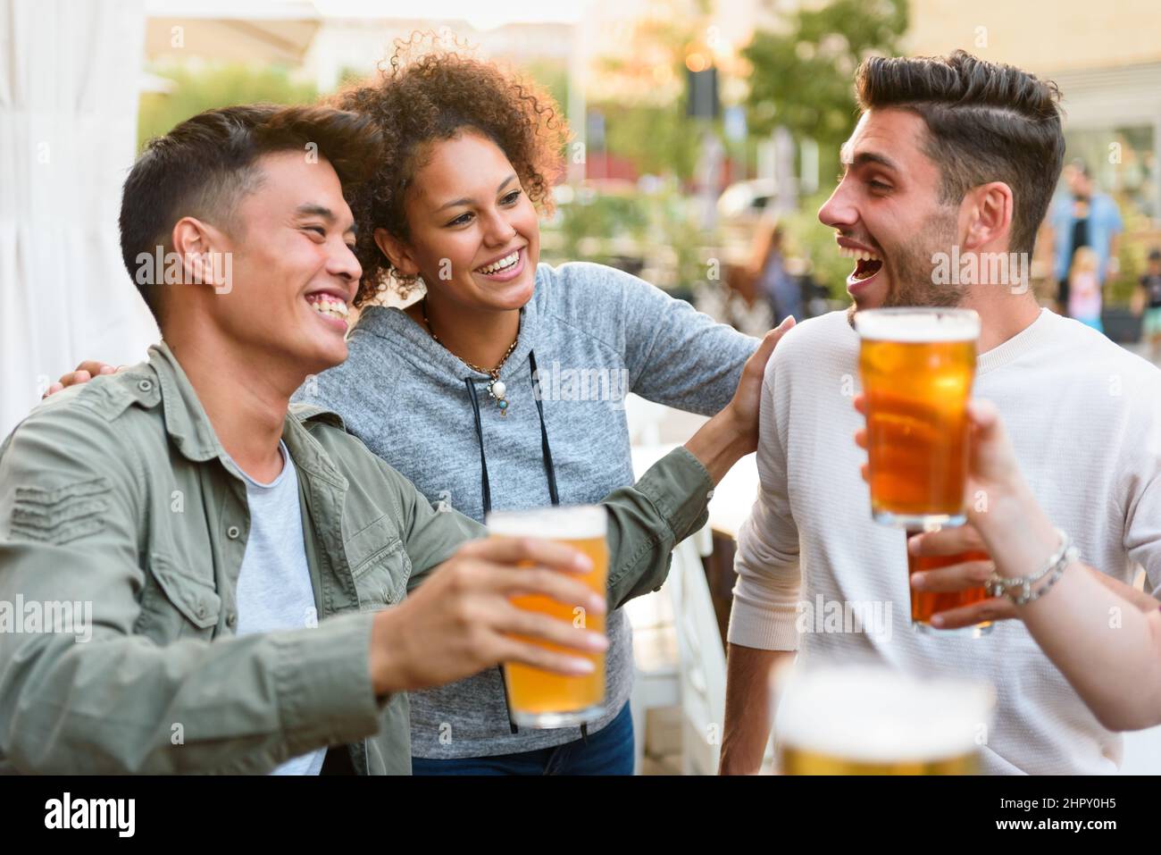 Gruppe von fröhlichen multirassischen Freunden, die einander beim Trinken von alkoholischem Bier auf der Terrasse des Cafés im Freien in der Stadt betrachten Stockfoto