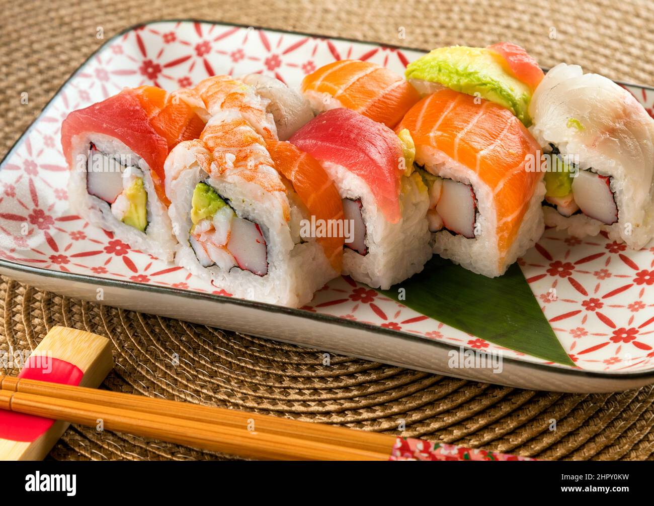 Appetitliche Uramaki-Sushi-Rollen in Regenbogenfarben mit Krabben und Avocado, gekrönt von verschiedenen Fischsorten, die in der Nähe von Essstäbchen auf dem Teller serviert werden Stockfoto