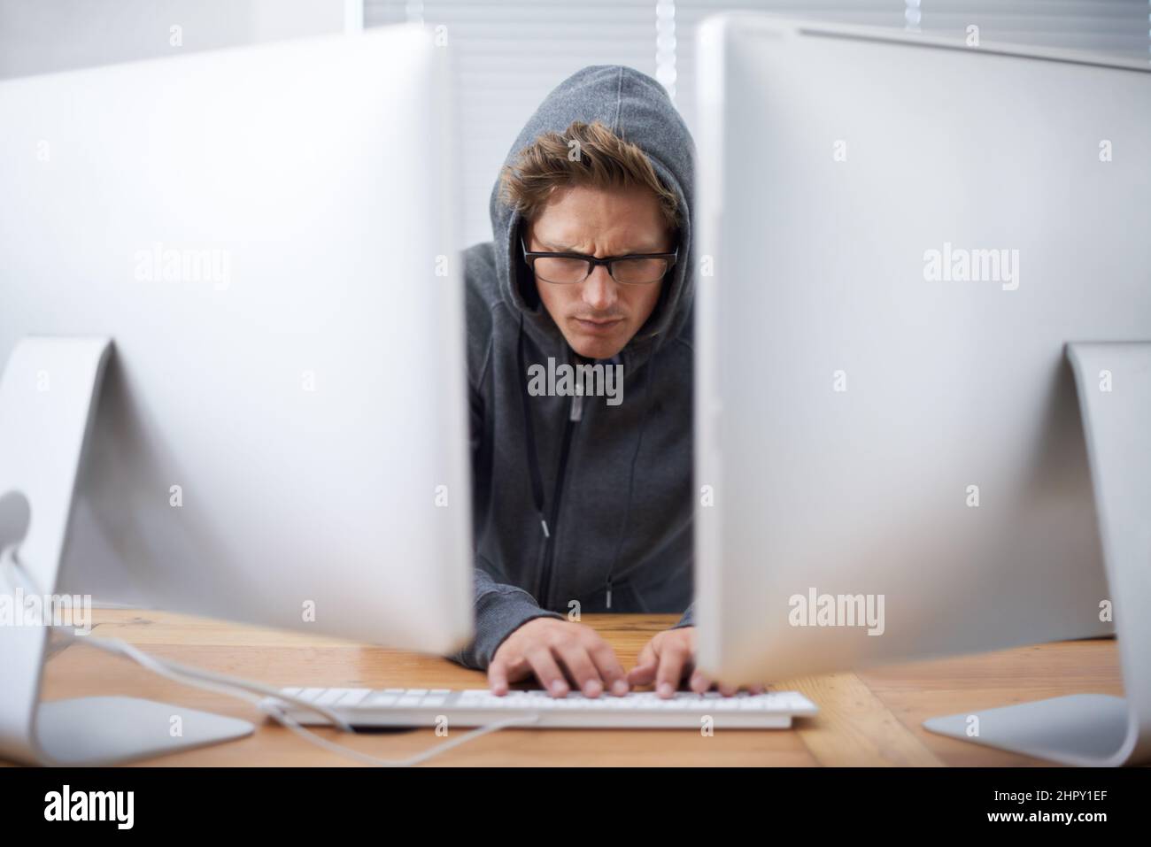 Das Mitternachtsöl verbrennen. Ein junger Hacker, der auf zwei Bildschirmen und einem Laptop mit Hoodie arbeitet. Stockfoto