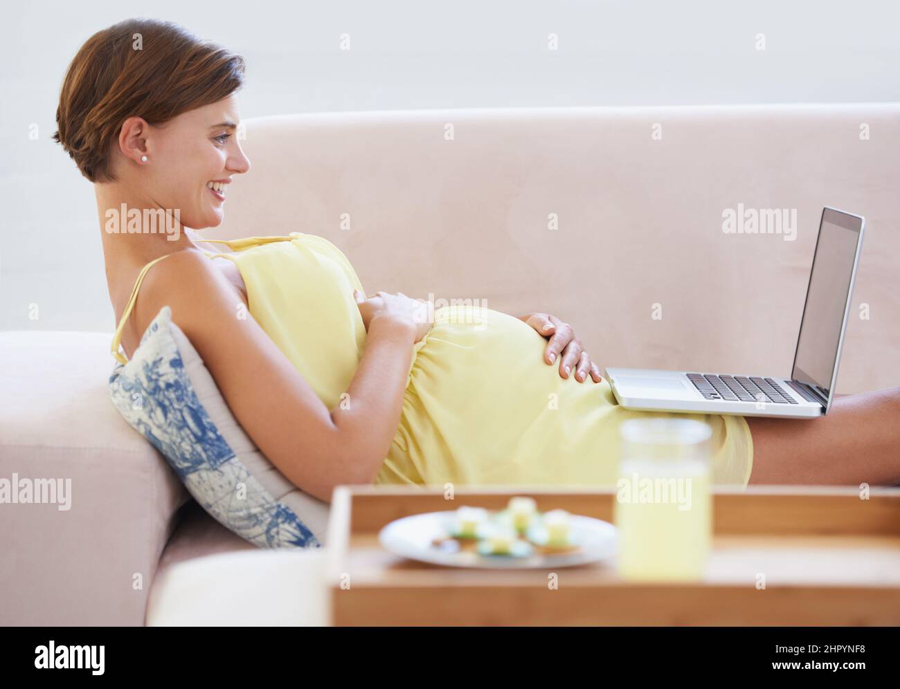 Gut gelaunt. Eine lächelnde Schwangerin, die mit einem Laptop auf der Couch liegt und neben ihr eine gesunde Mahlzeit serviert. Stockfoto