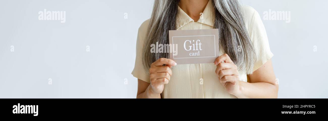 Lächelnde grauhaarige Asiatin hält den Geschenkgutschein auf hellem Hintergrund Stockfoto