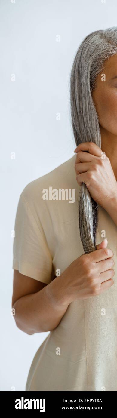 Entspannte asiatische Dame mittleren Alters in Bluse streicht Sperre von silbernen Haaren auf hellem Hintergrund Stockfoto