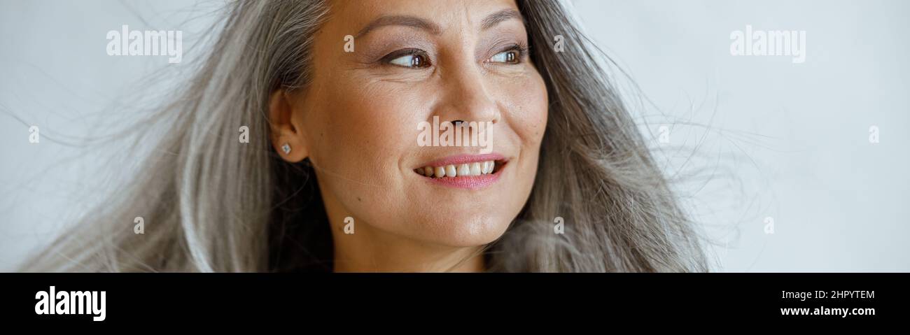 Glückliche asiatische Frau mittleren Alters mit fliegenden grauen Schlössern steht auf hellem Hintergrund Stockfoto