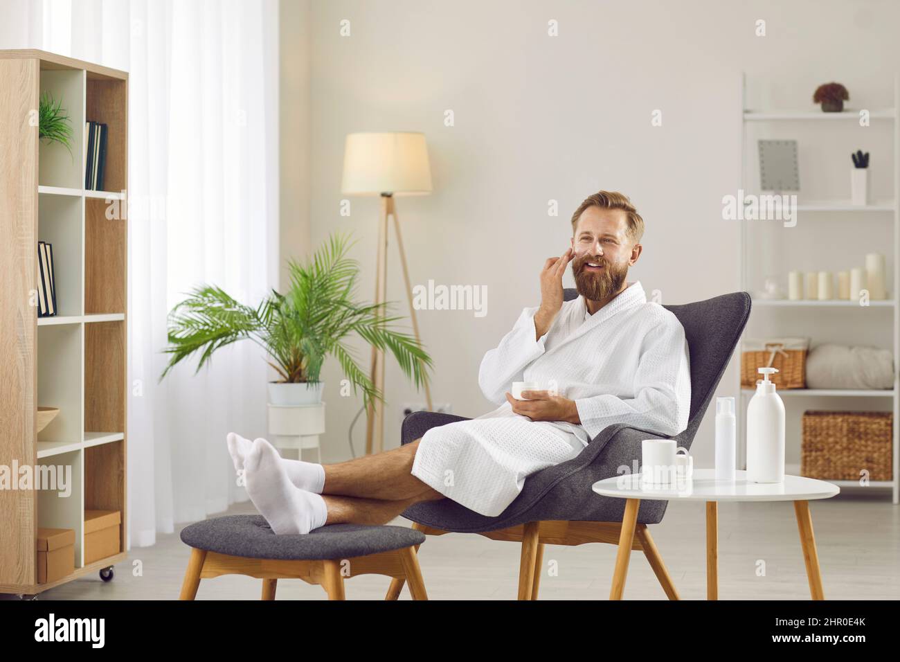 Glücklicher Mann im weißen Bademantel, der sich im Sessel entspannt und den Tag der Schönheit zu Hause genießt Stockfoto