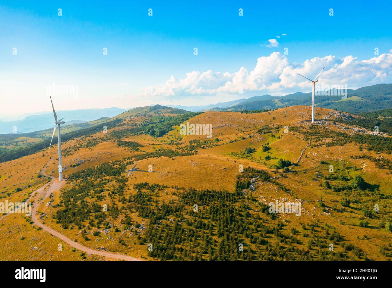 Windturbinen erzeugen alternative saubere Energie, die auf Hügelgipfeln mit Büschen im Hochland gegen forstwirtschaftliche Berge steht, bei sonniger Tagesansicht aus der Luft Stockfoto