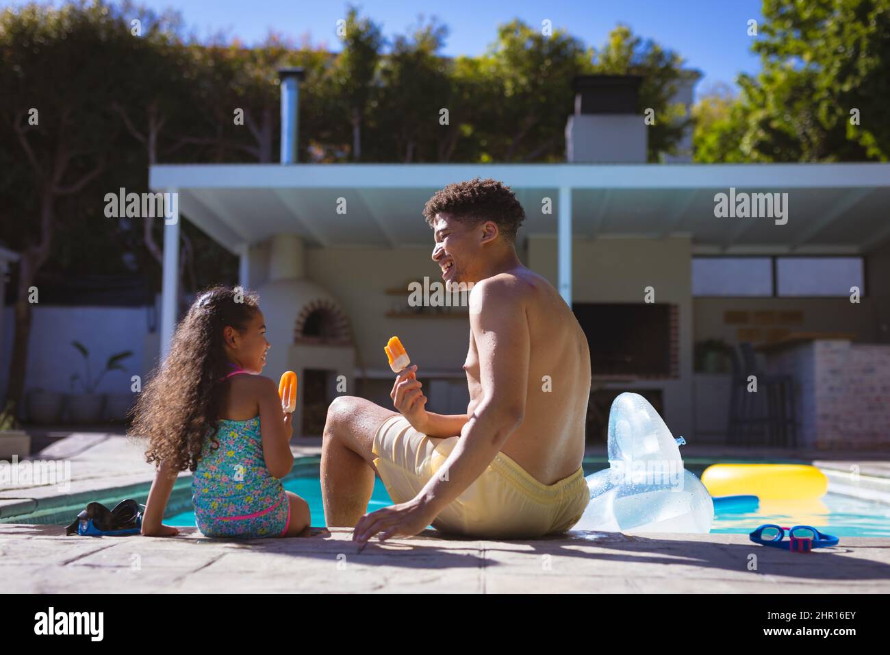 Multirassische Vater und Tochter essen aromatisierte Eisknolle, während sie zusammen am Pool sitzen Stockfoto