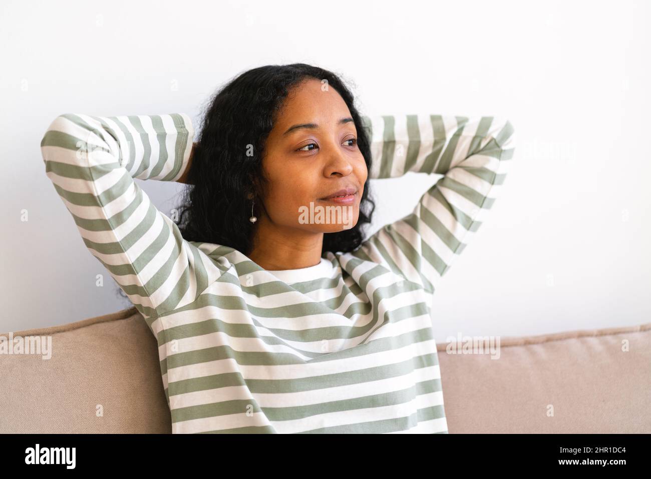 Glückliche afroamerikanische Frau, die die Hände hinter den Kopf legt, während sie im Wohnzimmer auf dem Sofa sitzt Stockfoto