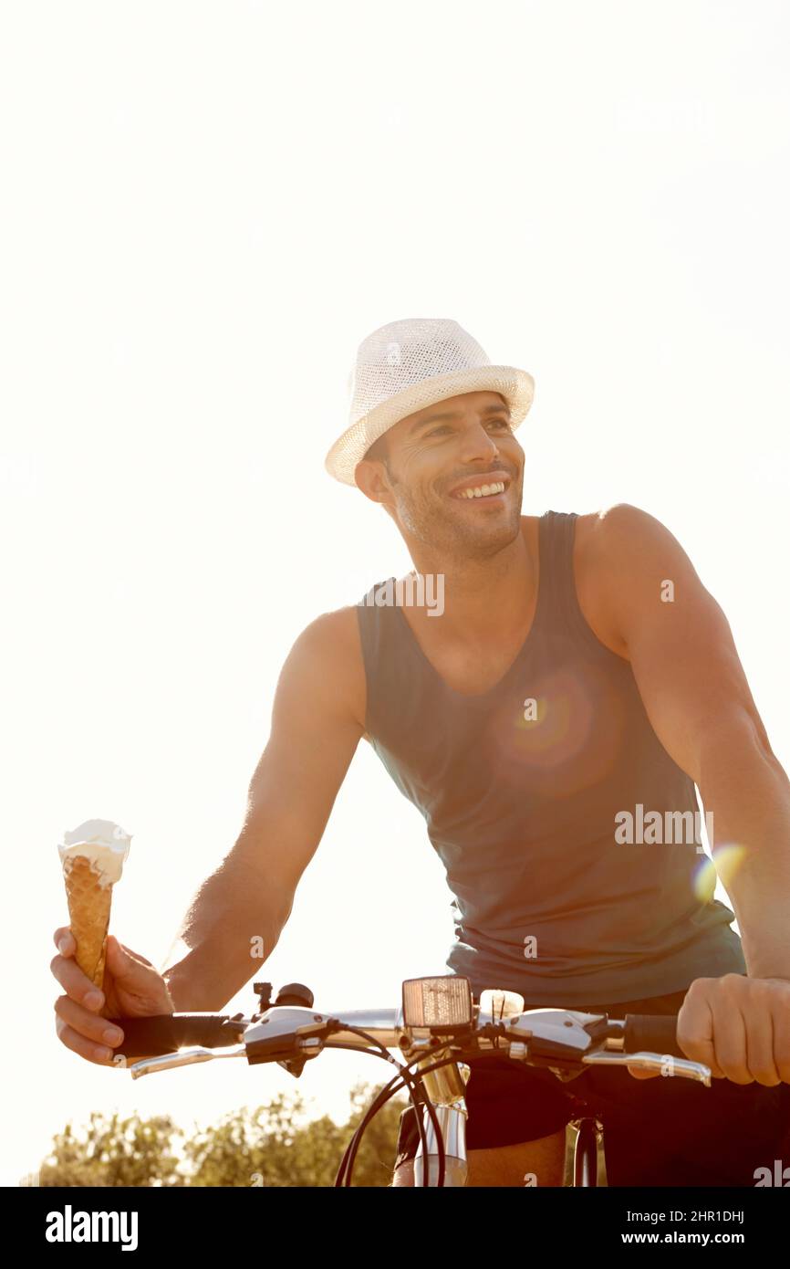 Genießen Sie einen Eiskegel und eine Radtour. Aufnahme eines hübschen jungen Mannes, der ein Eis isst, während er mit dem Fahrrad im Freien fährt. Stockfoto