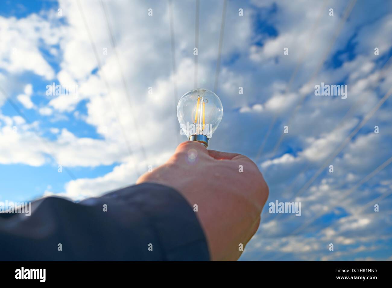 Strom und grüne Energie.Glühbirne in der Hand auf Stromleitung Hintergrund.Stromleitung Stromleitungen auf blauem Himmel Hintergrund.Alternative Stockfoto