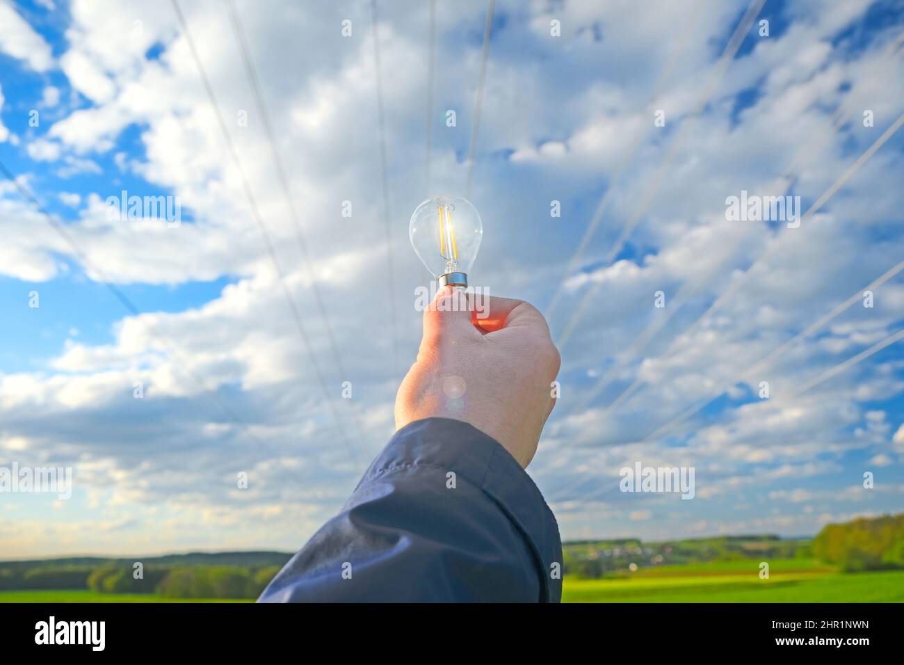 Strom und grüne Energie.Glühbirne in der Hand auf Stromleitung Hintergrund.Stromleitung Stromleitungen auf blauem Himmel und grünem Feld Stockfoto