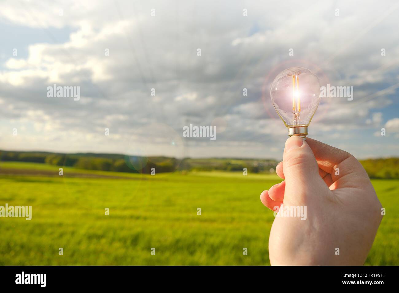 Strom und grüne Energie.Glühbirne in der Hand auf Stromleitung Hintergrund. Stromleitungen auf blauem Himmel und grünem Feld Hintergrund.Alternative Stockfoto