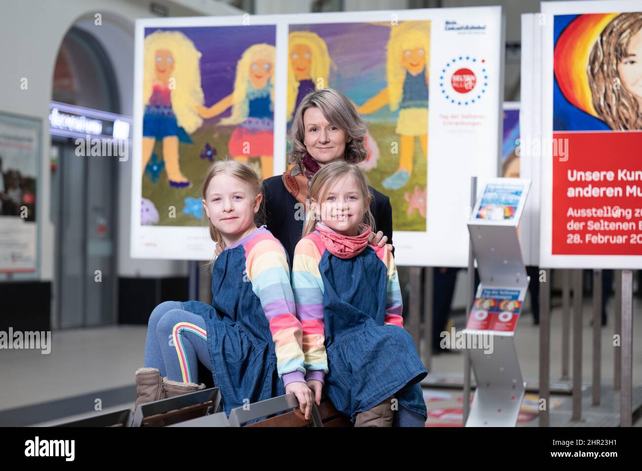 18. Februar 2022, Sachsen, Dresden: Emma (l.) und Lotte sitzen in einer Ausstellung zum Tag der Seltenen Erkrankungen vor dem Selbstporträt ihrer Mutter Katja Franke im Dresdner Hauptbahnhof. Die Zwillinge leiden an einer Neurofibromatose vom Typ 1, einer unheilbaren Krankheit. Mehr als vier Millionen Menschen in Deutschland leiden an einer von mehr als 6.000 Krankheiten, die nur sehr wenige andere haben. Der Welttag der Seltenen Krankheiten erinnert an die Sorgen der Betroffenen am 28. Februar 2022. Foto: Sebastian Kahnert/dpa-Zentralbild/dpa - ACHTUNG: Nur für redaktionelle Verwendung im Zusammenhang mit der aktuellen Berichterstattung über die Seltene Krankheiten Da Stockfoto