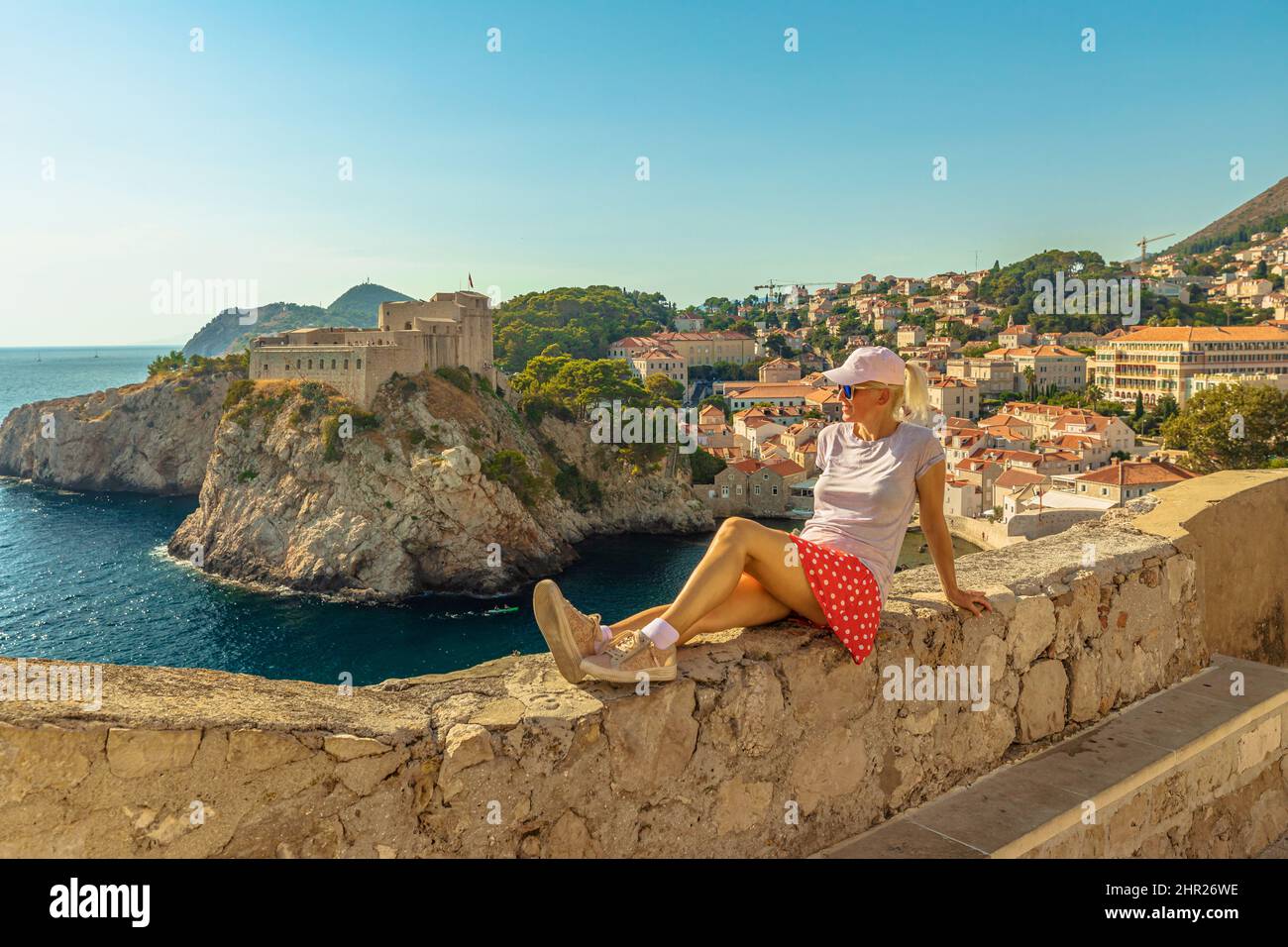 Frau, die auf den oberen Wänden der Stadt Dubrovnik in Kroatien sitzt. Blick auf die Festung Fort Lovrijenac, über den Westhafen. Dubrovnik historische Stadt von Kroatien Stockfoto